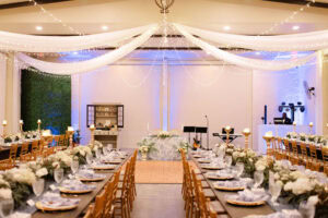 Elegant wedding reception at Bonita Ranch with long wooden guest tables, French toile napkins, white garden florals, gold chairs, candlelight, drapery, fairy lights, sweetheart table, and DJ setup.