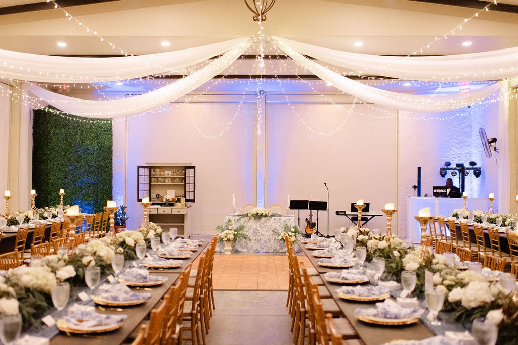 Elegant wedding reception at Bonita Ranch with long wooden guest tables, French toile napkins, white garden florals, gold chairs, candlelight, drapery, fairy lights, sweetheart table, and DJ setup.