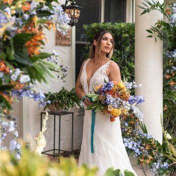 A bride posing under the Spanish porch at Bonita Ranch, with columns beautifully decorated with colorful flowers by Fern and Curl Florist, captured by Winship Photography.
